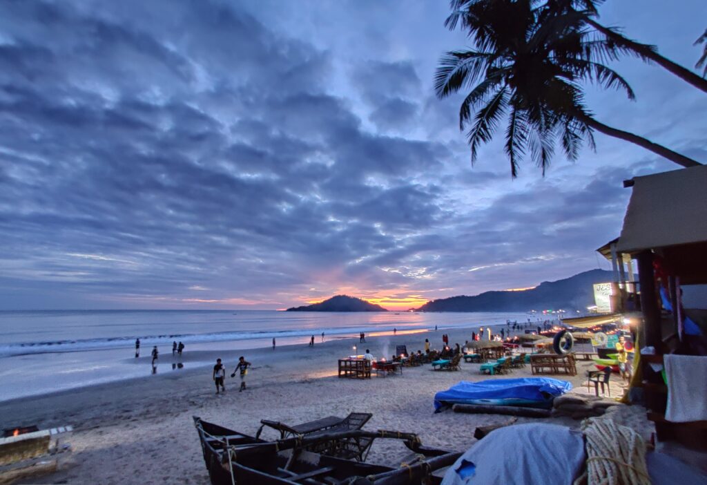 A beach at sunset with several people sitting on the sand, enjoying the view of the colorful sky and ocean waves.